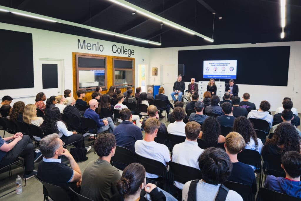 A large group of students and community members sit facing a panel of four speakers in a Menlo College classroom during an ‘AI and Sustainability’ discussion. The speakers sit on a stage in front of a screen displaying the event title, while the audience fills the room.